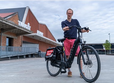 Beschrijving afbeelding: jongeman toont trots zijn aangepaste elektrische fiets. Hij staat op een plein met op de achtergrond een hellend vlak richting inkom van een gebouw. Op het gebouw de letters TRANZIT en DEPART.