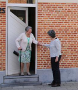 Beschrijving afbeelding: oudere vrouw met steunstok staat in haar geopende deur en schudt de hand van een jongere dame op straat.