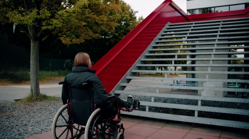 Beschrijving afbeelding: vrouw in rolstoel kijkt tegen een enorme trappenpartij op.