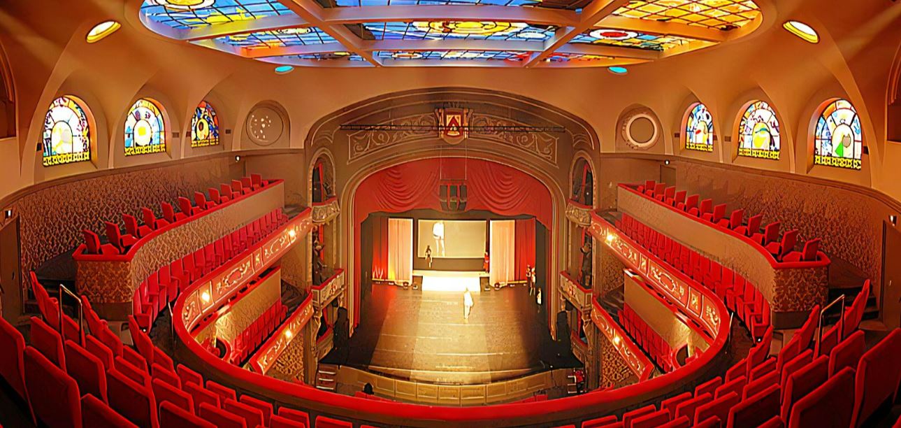 Beschrijving afbeelding: binnenzicht van de Kortrijkse schouwburg met het kleurrijk glas in lood plafond en de rode pluche zeteltjes in arenavorm.