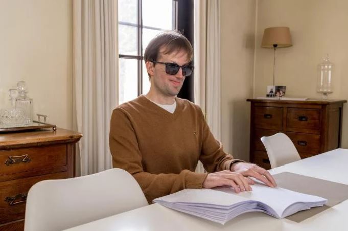 Jongeman zit aan tafel, draagt een bril met donkere glazen, hij leest braille in een geopend boek dat voor hem ligt op tafel