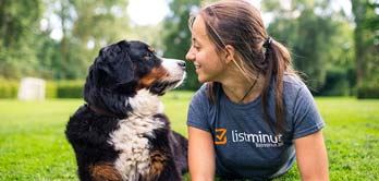 Een jonge vrouw kijkt liefdevol in de ogen van een Berner sennenhond. Zij liggen naast elkaar op een grasplein. De vrouw draagt een t-shirt met de naam en het logo van listminut. De hoofdkleur van de grote hond is zwart, verder is er wit bij de snuit, hal