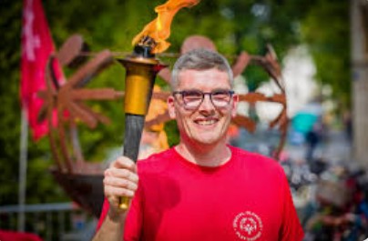 Beschrijving afbeelding: man in rode t-shirt draagt in zijn rechter hand de brandende Olympische fakkel, op zijn gezicht een brede glimlach.