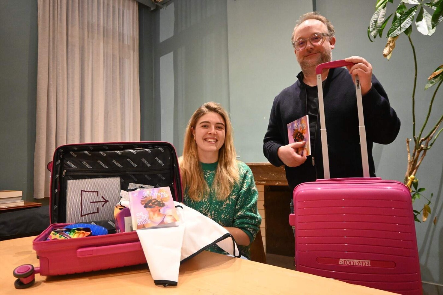 Beschrijving afbeelding: twee lachende jonge mensen aan tafel met flashy reiskoffers bij de hand.