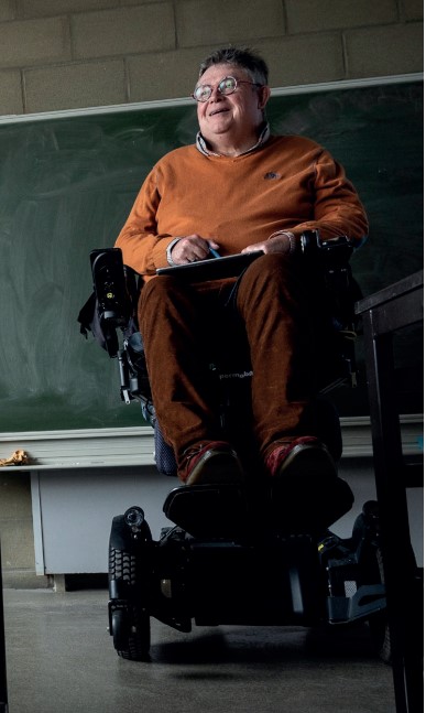 Beschrijving afbeelding: rolstoelgebruiker Chris, breed glimlachend, heeft zich gepositioneerd voor een bord in een klaslokaal.